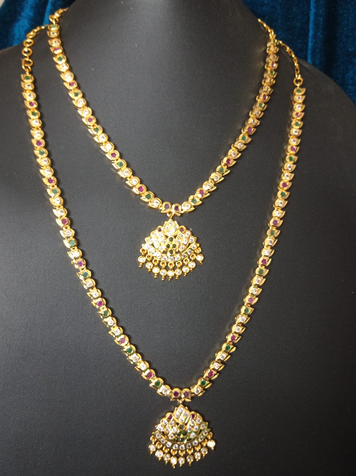 Pure Panchaloham two-layer haram with gold-tone detailing, red and green stones, clear accents, and traditional dome-shaped dangling pendants displayed on a black jewellery bust.
